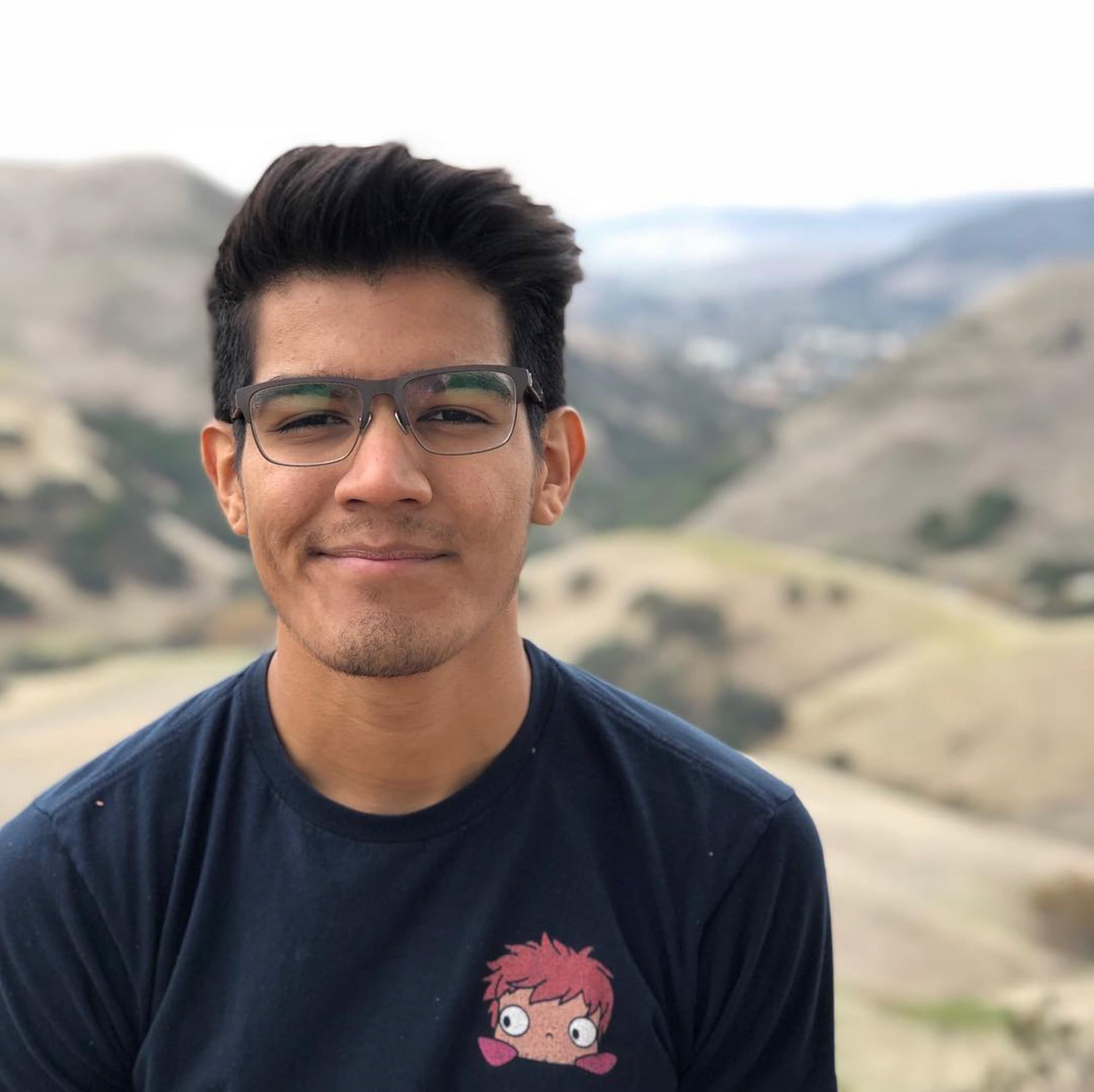 Antonio on a ridge above the San Luis Obispo — dark short hair swept up, square frames, a navy tee with a small Ponyo print, hills out of focus behind.